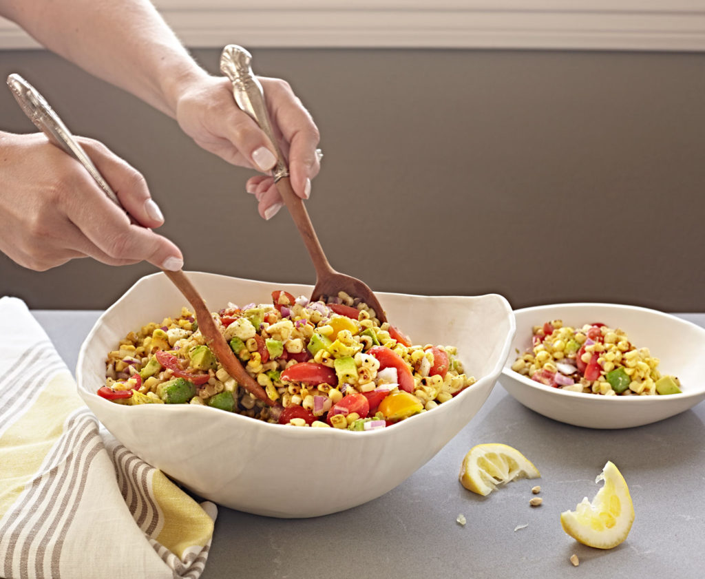 Hands tossing summer Corn Salad with tomatoes and avocado