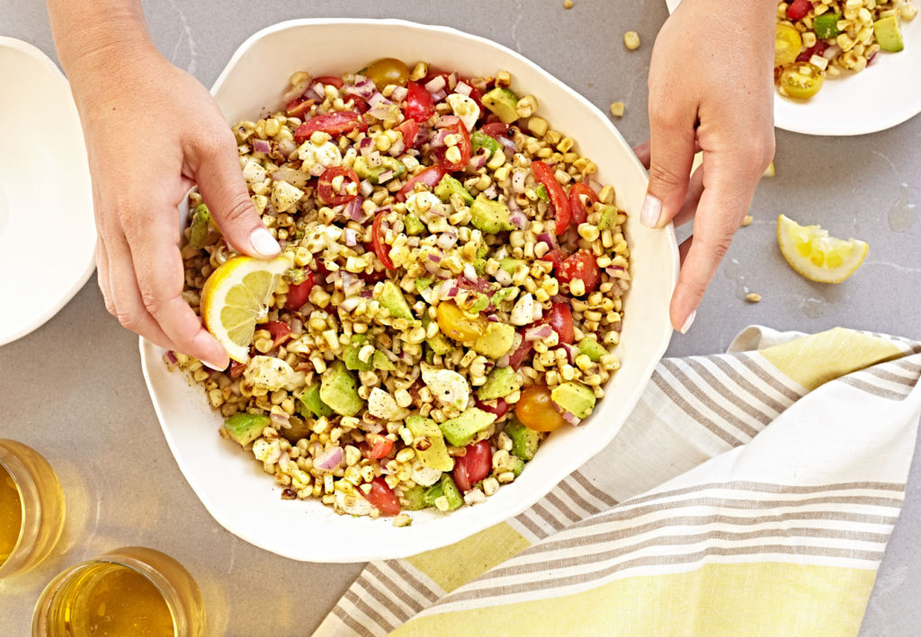 hands squeezing lemon into summer Corn Salad with tomatoes and avocado