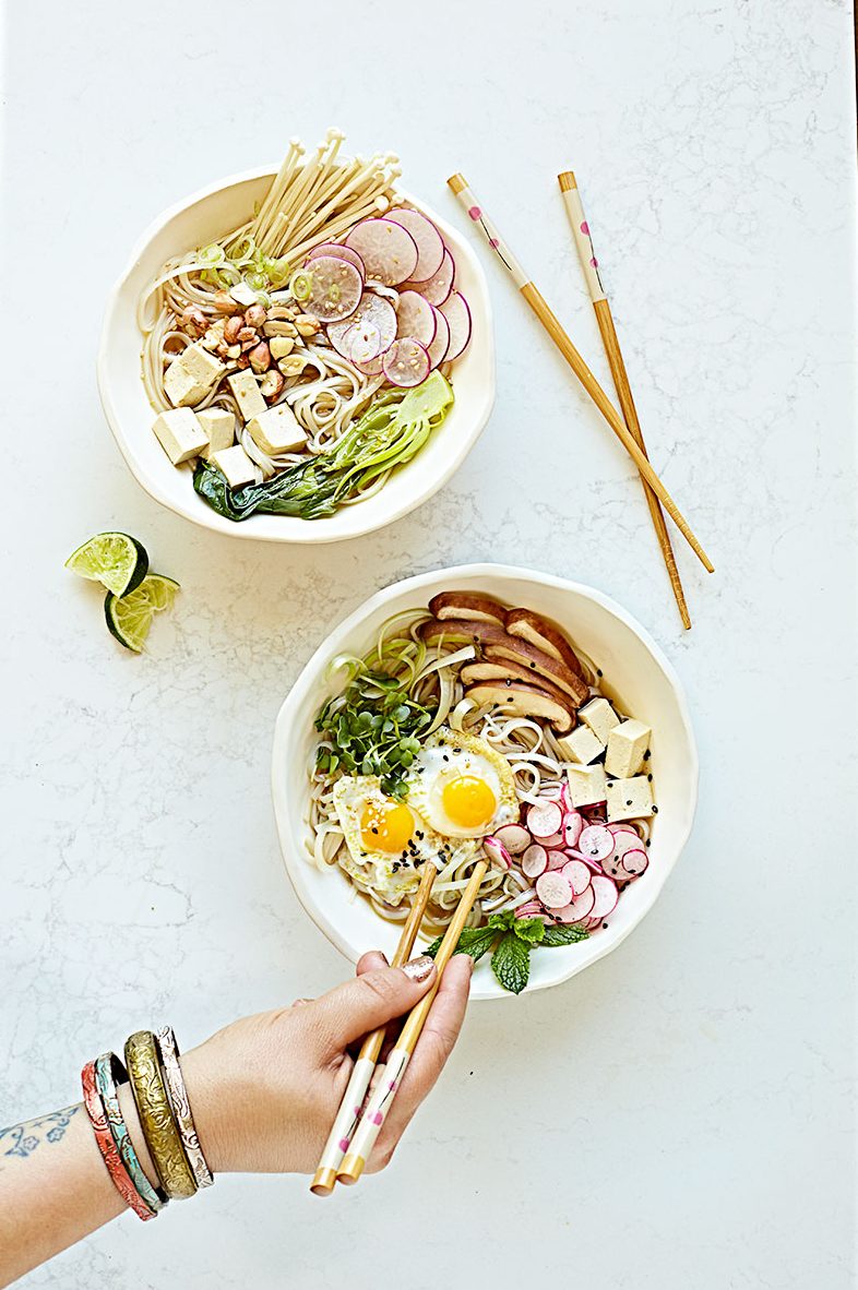 Two bowls of vegetarian pho noodle soup with different toppings and a hand reaching in to eat it.
