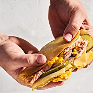 Man's hands holding a pressed gluten-free crepe cuban sandwich with ham, pork, mustard and pickles.